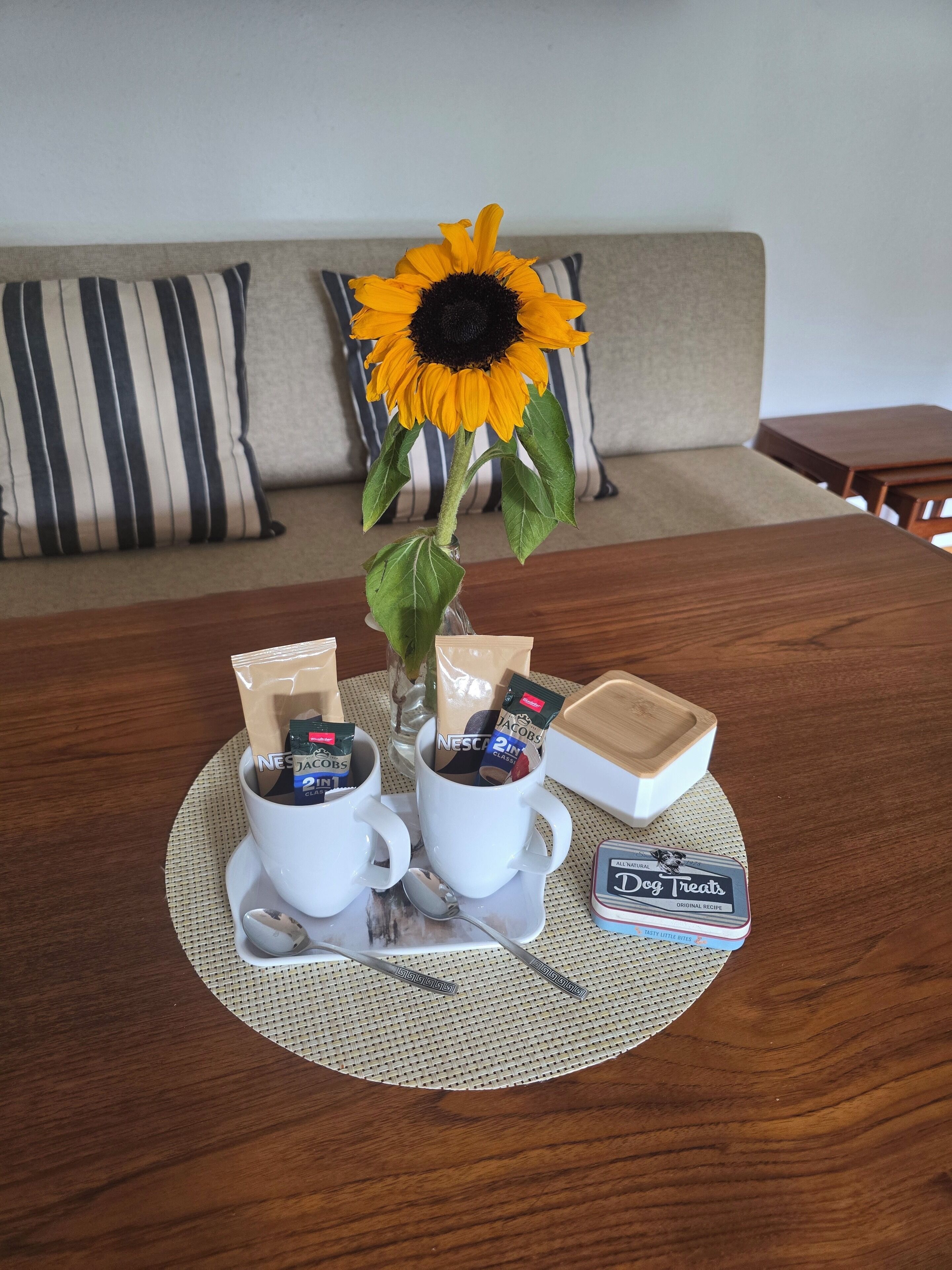 A round wooden table with two white coffee cups, a vase with a sunflower, and a box of dog treats.