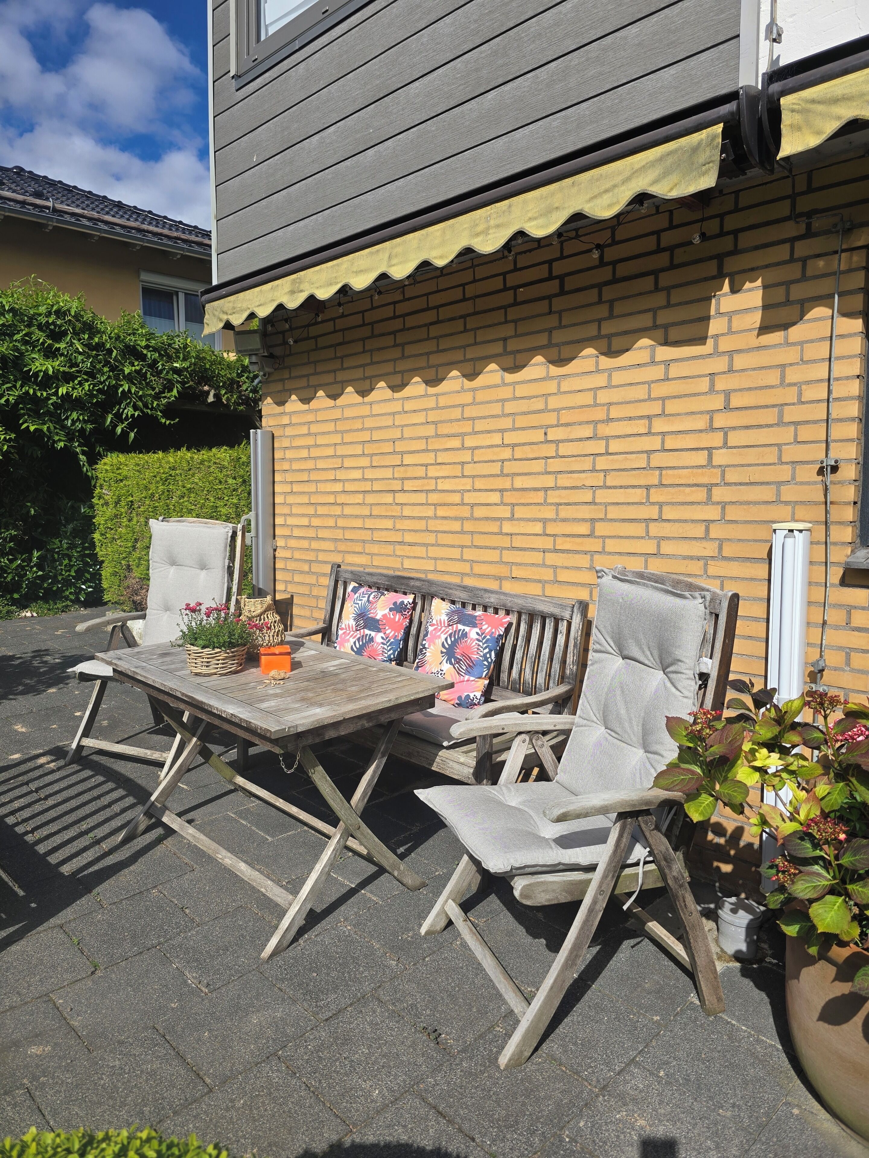 Une terrasse extérieure avec une table et des chaises en bois, un mur en briques et un auvent jaune.