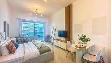 A modern hotel room with a bed, a dining area, a TV, and a view of the city.
