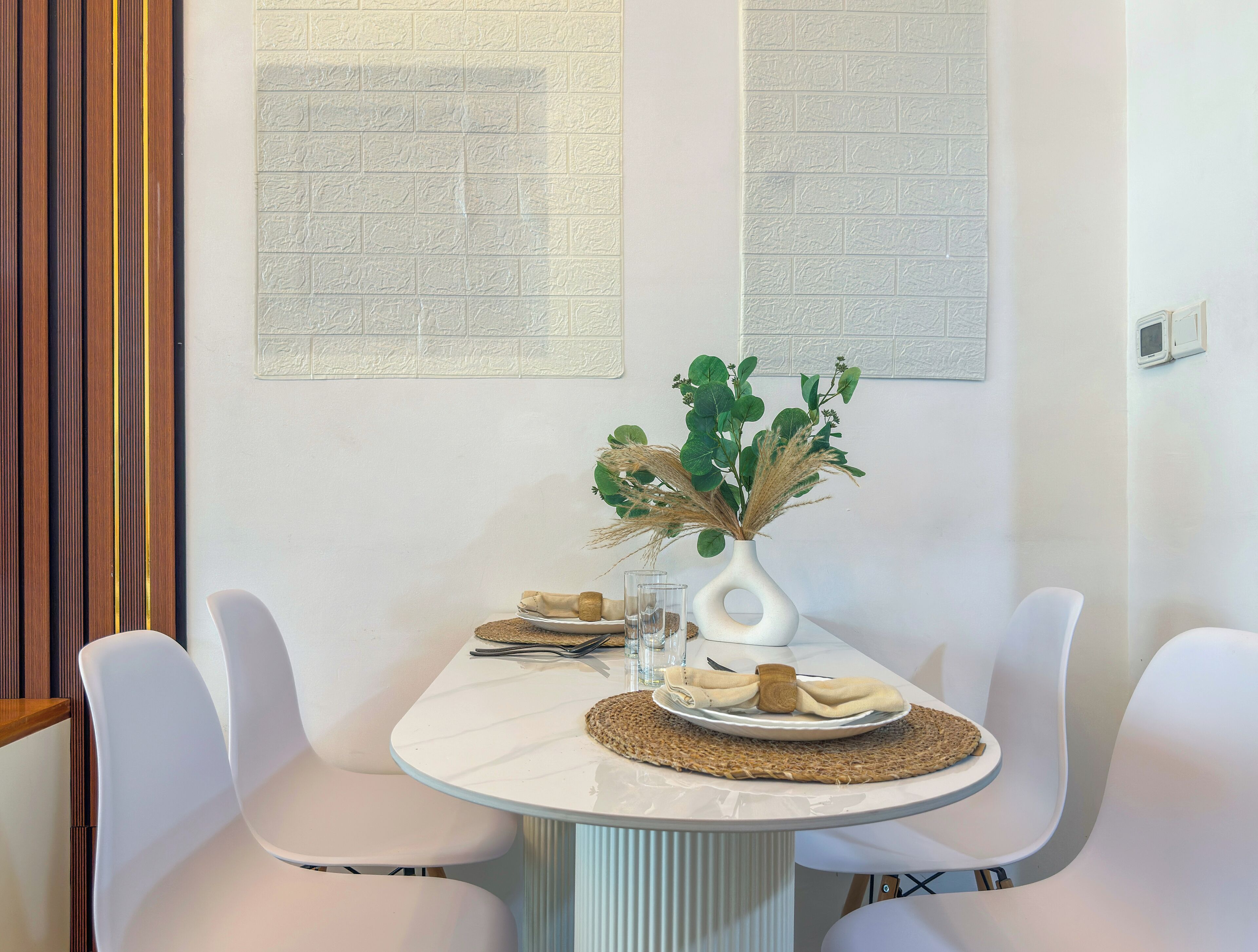 A dining area with a white table, two white chairs, a vase with greenery, a plate with bread, and a textured wall.