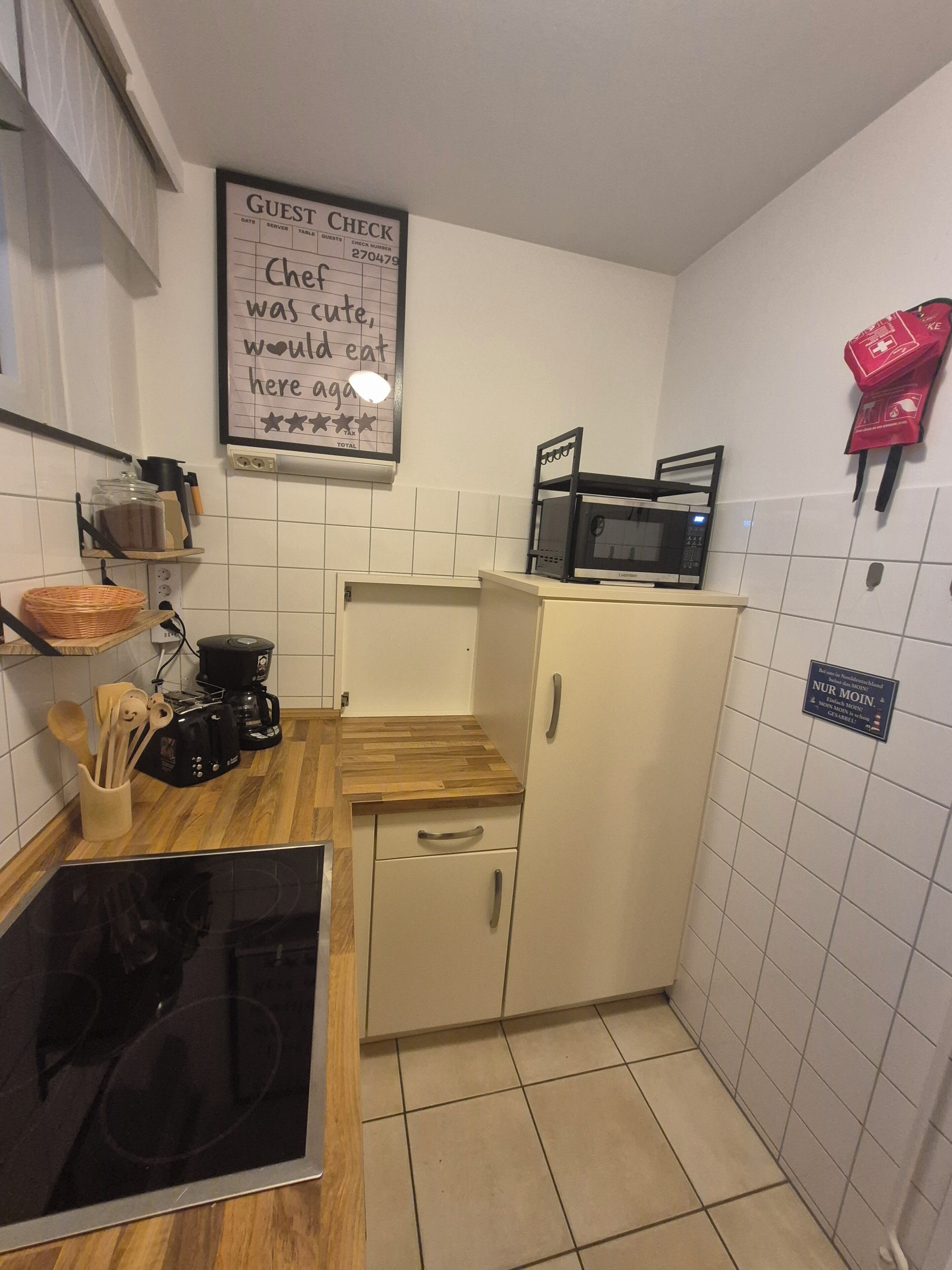 Uma pequena cozinha compacta com micro-ondas, cafeteira e uma prateleira com utensílios de cozinha. Há um aviso na parede e uma bolsa vermelha pendurada.