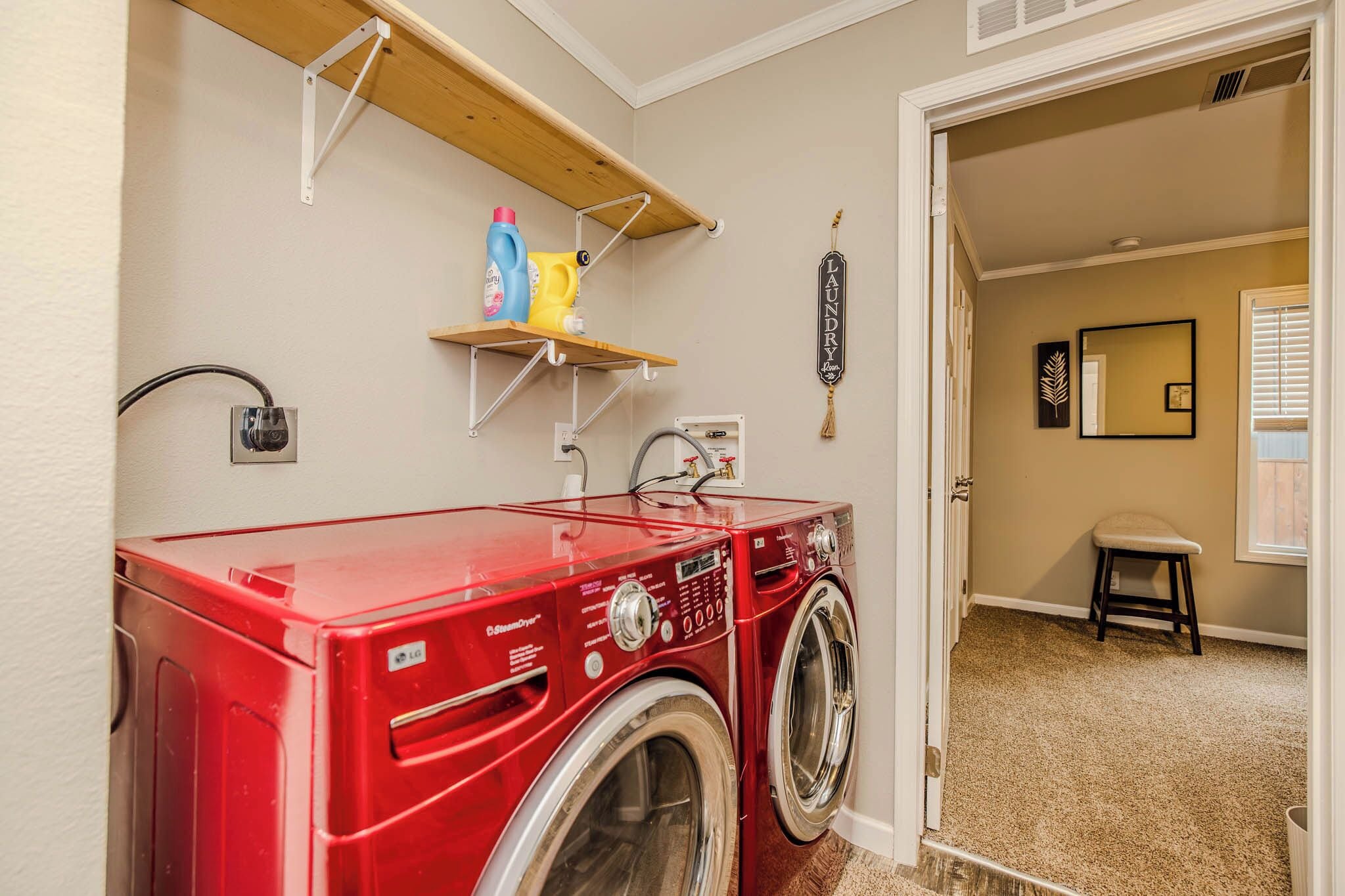 Une buanderie avec un lave-linge et un sèche-linge à chargement frontal de couleur rouge, une étagère contenant des produits de lessive et une porte donnant sur une autre pièce.