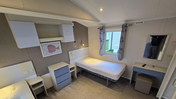 Une pièce de taille modeste comprenant un lit, un petit bureau, une table de chevet, un miroir et une fenêtre avec des rideaux.