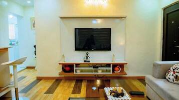 Una sala de estar con una televisión de pantalla plana montada sobre un soporte de madera, una mesa de centro con objetos decorativos y un sofá con un cojín estampado.