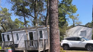 A mobile home with a wooden deck, a car parked nearby, and a tree in the foreground. - Place of Peace, (Camiers)