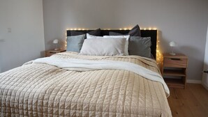 Un lit bien fait, recouvert d’une couette beige, avec deux tables de chevet dotées de lampes et une tête de lit décorée de guirlandes lumineuses.