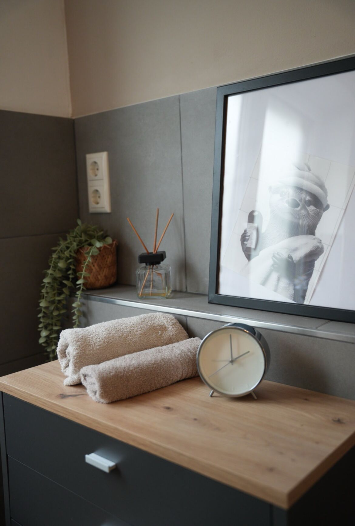 Un bagno ben organizzato con un mobile bagno in legno, un quadro incorniciato, una pianta in vaso e una piccola sveglia.