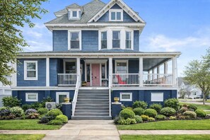 A two-story house with a front porch, a covered front porch area, and a front yard with various plants. - 10BR North WW Beach Home - Sleeps 22! (North Wildwood)