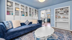 Een woonkamer met een blauwe bank, een witte salontafel en een kamerbreed tapijt met een patroon. Er zijn witte kasten met glazen deuren en een witte plank met decoratieve objecten.