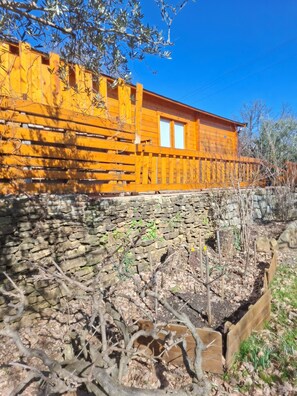 Un bâtiment rustique en bois, doté de fondations en pierre, entouré d’une végétation aride et surplombé d’un ciel bleu clair.