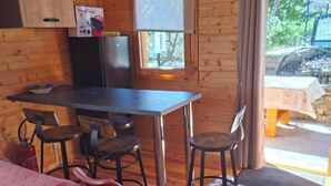 A wooden cabin interior with a dining table, chairs, a kitchen area, and a window with curtains. - CHALET MARYVONNE LES VANS 07140 (Les Vans)