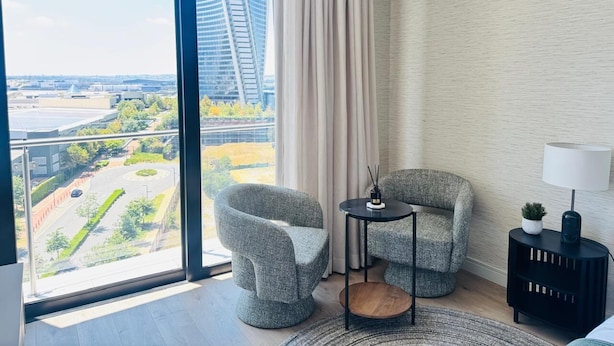 Ett modernt hotellrum med ett stort fönster som erbjuder utsikt över staden, två stoppade fåtöljer, ett litet runt bord, en lampa och ett sidobord med en växt.