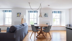 Ein modernes Wohnzimmer mit einer Couch, einem Esstisch und Stühlen. Große Fenster mit blauen Vorhängen, ein Heizkörper und Kunstwerke an den Wänden.