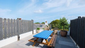 Eine Terrasse im Freien mit einem Holztisch und Bänken, umgeben von einem Holzzaun und Blumentöpfen.