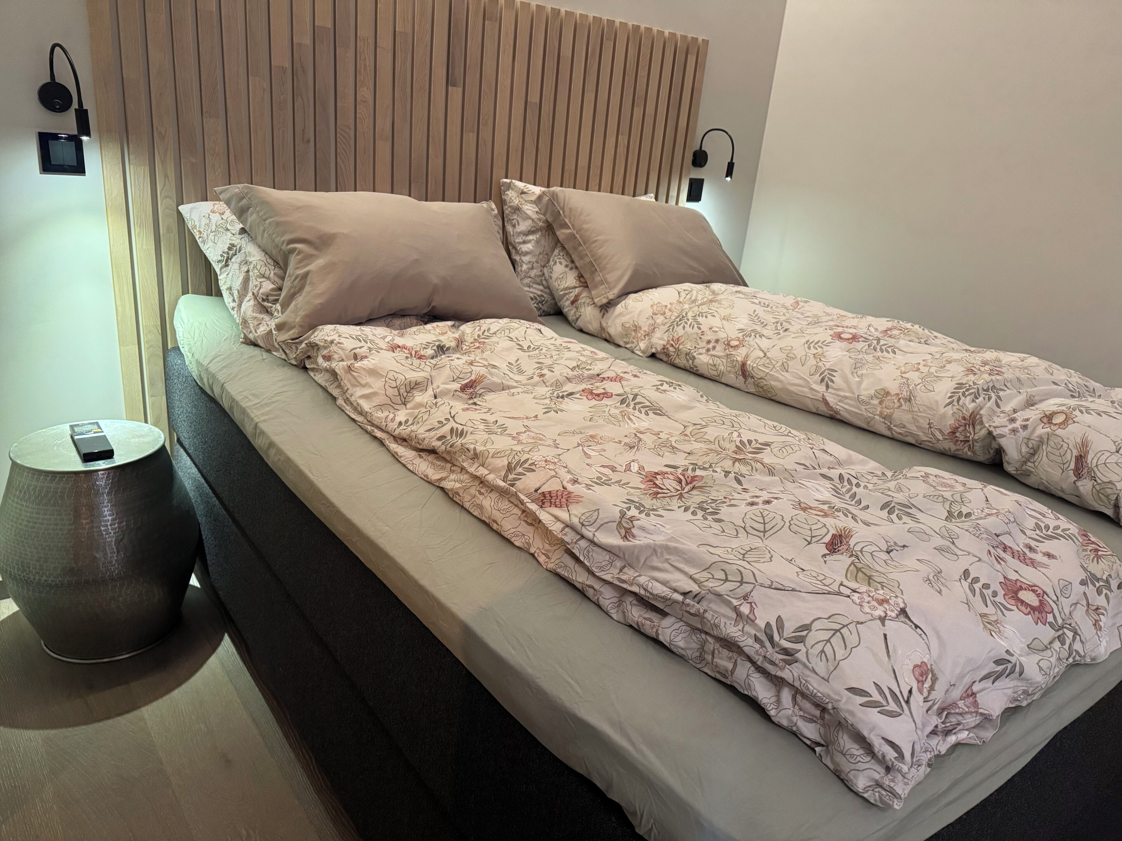 A single bed with a floral bedspread, two pillows, a bedside lamp, and a small round table with a device on it.