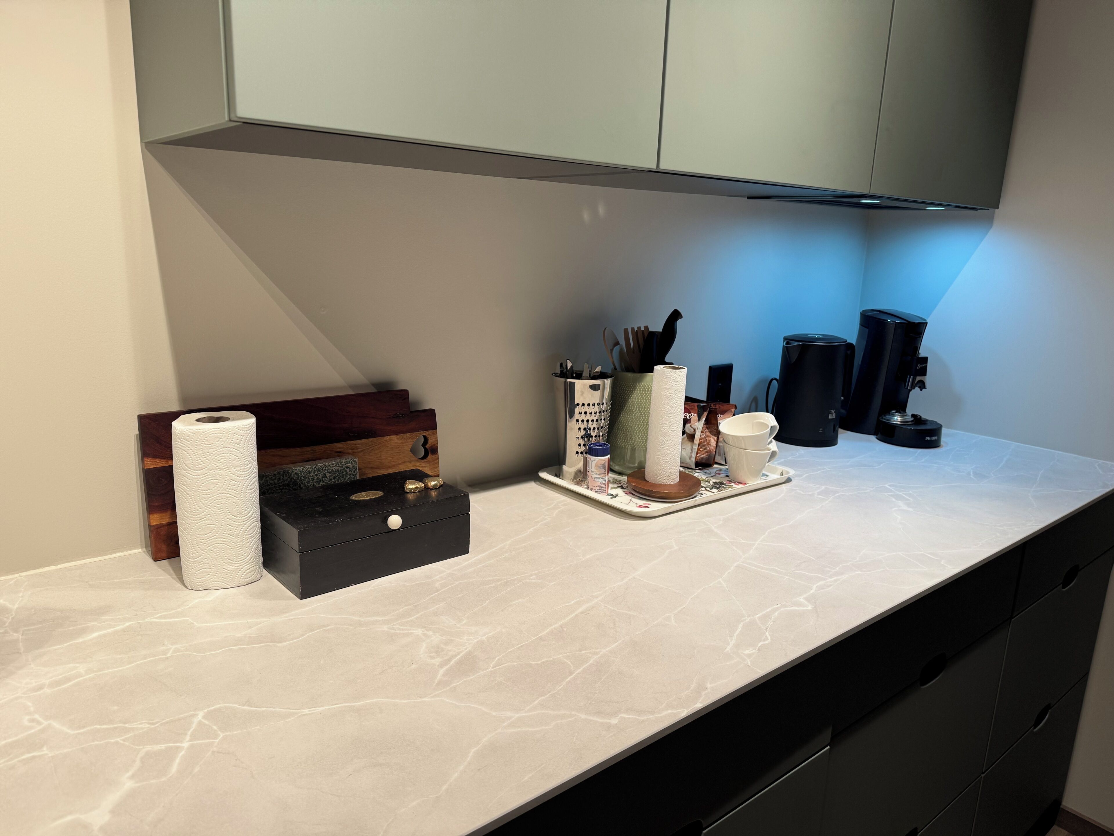 A kitchen counter with a paper towel dispenser, a spice rack, a coffee maker, and a tray with cups and a bottle.