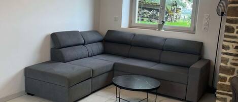 A modern living room with a sectional sofa, a small round table, and a large window with a view of greenery.