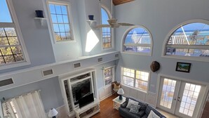 A spacious living room with a fireplace, large windows, and a high ceiling with wooden beams.