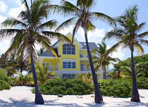 A two-story yellow building with a balcony, surrounded by palm trees and a sandy beach.