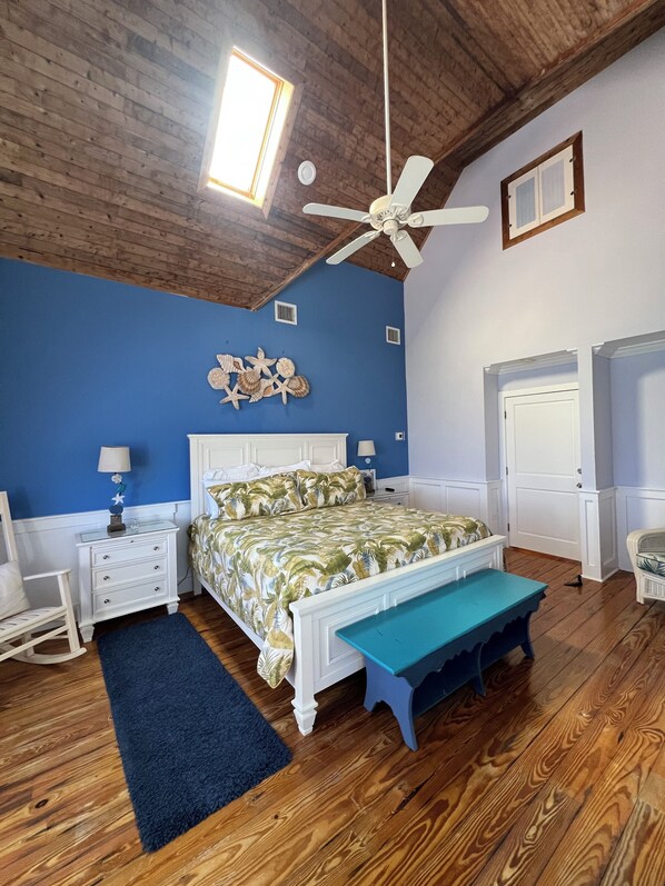 A bedroom with a wooden ceiling, a bed with a blue bench, a rocking chair, and a wall with seashell decorations.