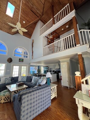 A spacious living area with a high ceiling, wooden beams, a large sofa, a dining table, and a kitchen in the background.