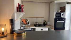 Una cocina compacta con estufa a gas, microondas y horno pequeño. Hay una estantería con utensilios de cocina y una vela sobre la encimera.