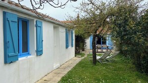 Filas de edificios blancos con contraventanas azules, un camino empedrado y un pequeño jardín con un árbol y mobiliario exterior.
