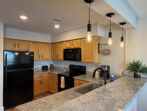A modern kitchen with granite countertops, wooden cabinets, a black refrigerator, a microwave, a stove, and pendant lighting.