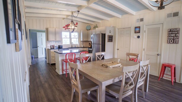 A kitchen with wooden cabinets, a refrigerator, and a dining area with a table and chairs. - Sweet Tee: Costal Cottage, Steps from the Beach (Ocean Isle Beach)