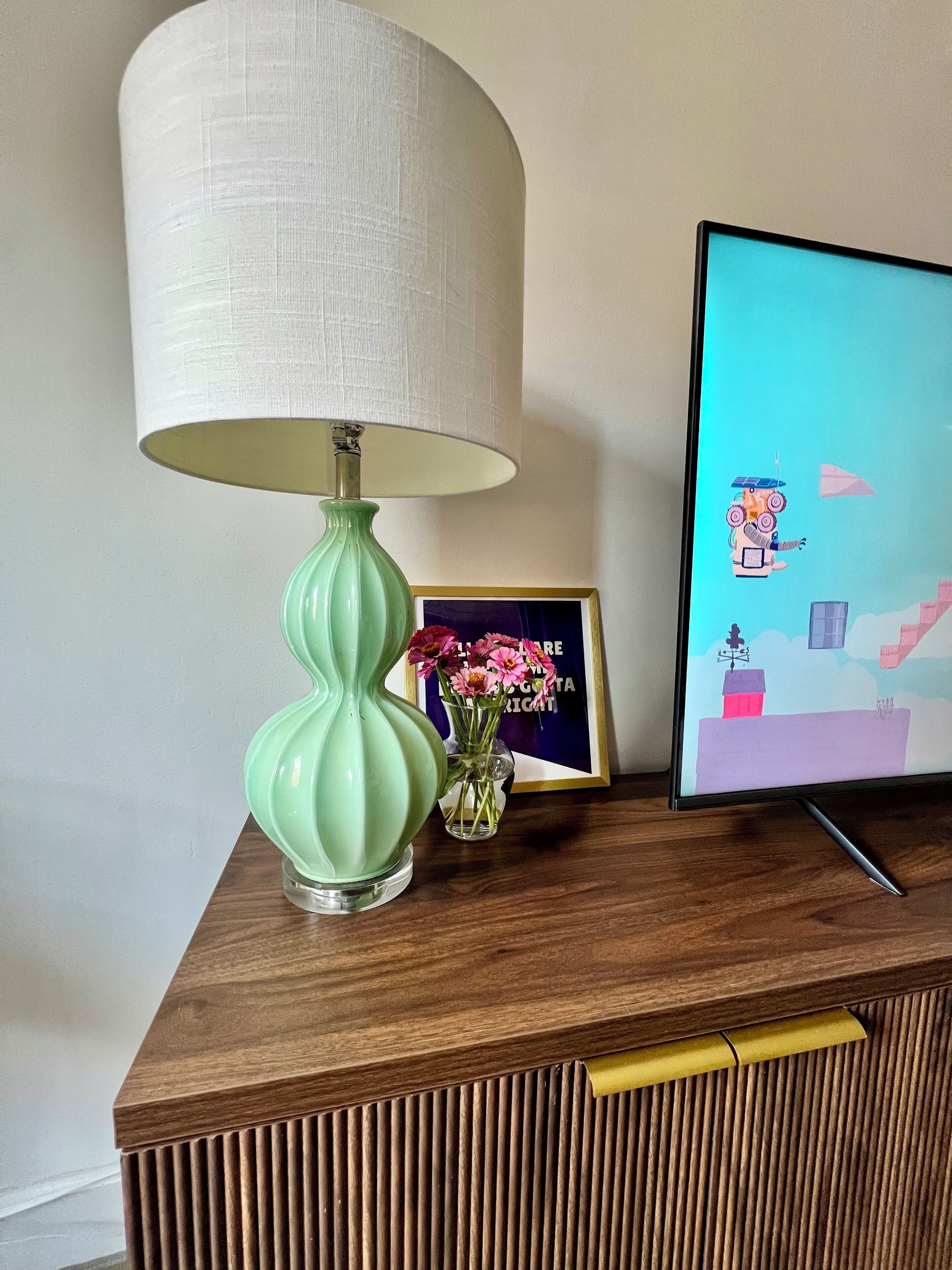 Ein Holzschrank mit einer grünen Lampe, einem gerahmten Bild und einer kleinen Vase mit Blumen.
