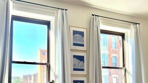 Ein Zimmer mit zwei großen Fenstern, einem schwarzen Sessel, einem kleinen Tisch mit Lampe und einem gemusterten Teppich.