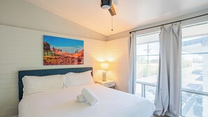 A hotel room with a large bed, a painting on the wall, a nightstand with a lamp, and a view through a window. - Dwell 7003-3 (Scottsdale)