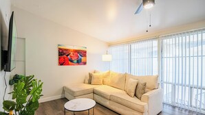 A living room with a beige sectional sofa, a round coffee table, a large window with blinds, a painting on the wall, and a potted plant. - Dwell 7003-3 (Scottsdale)