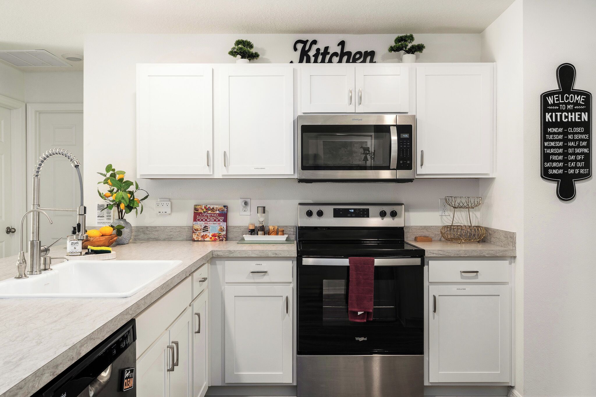 A modern kitchen with white cabinets, a stainless steel microwave, and an oven. There is a sign on the wall indicating the kitchen's operational hours.