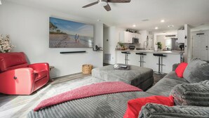 Ein modernes Wohnzimmer mit einer Couch, einem Hocker und einem roten Sessel, einem Küchenbereich mit Barhocker und einem Deckenventilator.