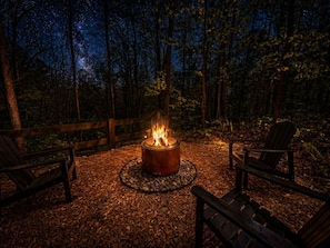 A campfire surrounded by wooden chairs and a circular stone fire pit in a forest. - The Overlook | Modern Cabin | Hot Tub | Fire Pit (Beattyville)