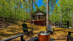 A cabin with a deck, surrounded by trees, and a fire pit with seating.