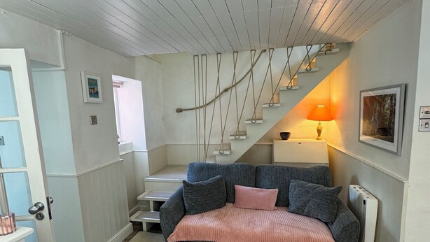 Een gezellige woonkamer met een grijze bank, een roze plaid en een houten salontafel. Een trap met een touwladdertje is op de achtergrond te zien.