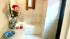 Een gang met een witte bank, een vaas met bloemen, een spiegel en een plafondlamp.