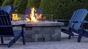 Un brasero rodeado de dos sillas Adirondack, ubicado en un patio empedrado con un seto al fondo.