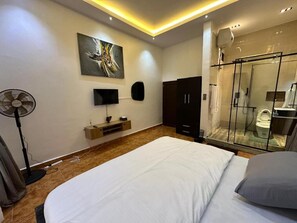 A modern bedroom with a large bed, a TV, a fan, and a glass-enclosed shower area. - BORA HOTEL (Ngwo)