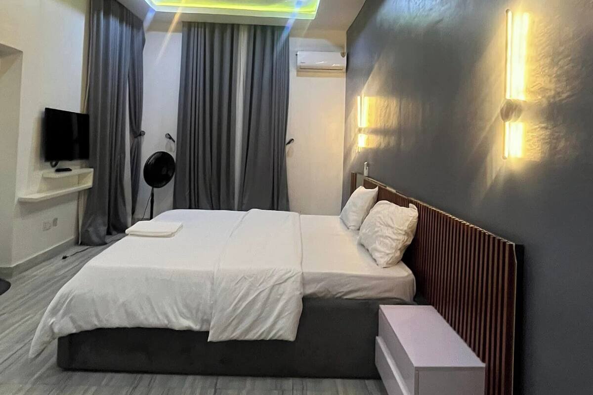 A modern hotel room with a large bed, a bedside table, a TV, and a ceiling light fixture.
