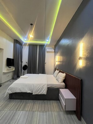 Quarto de hotel moderno com uma cama grande, mesa de cabeceira, televisão e luminária no teto.