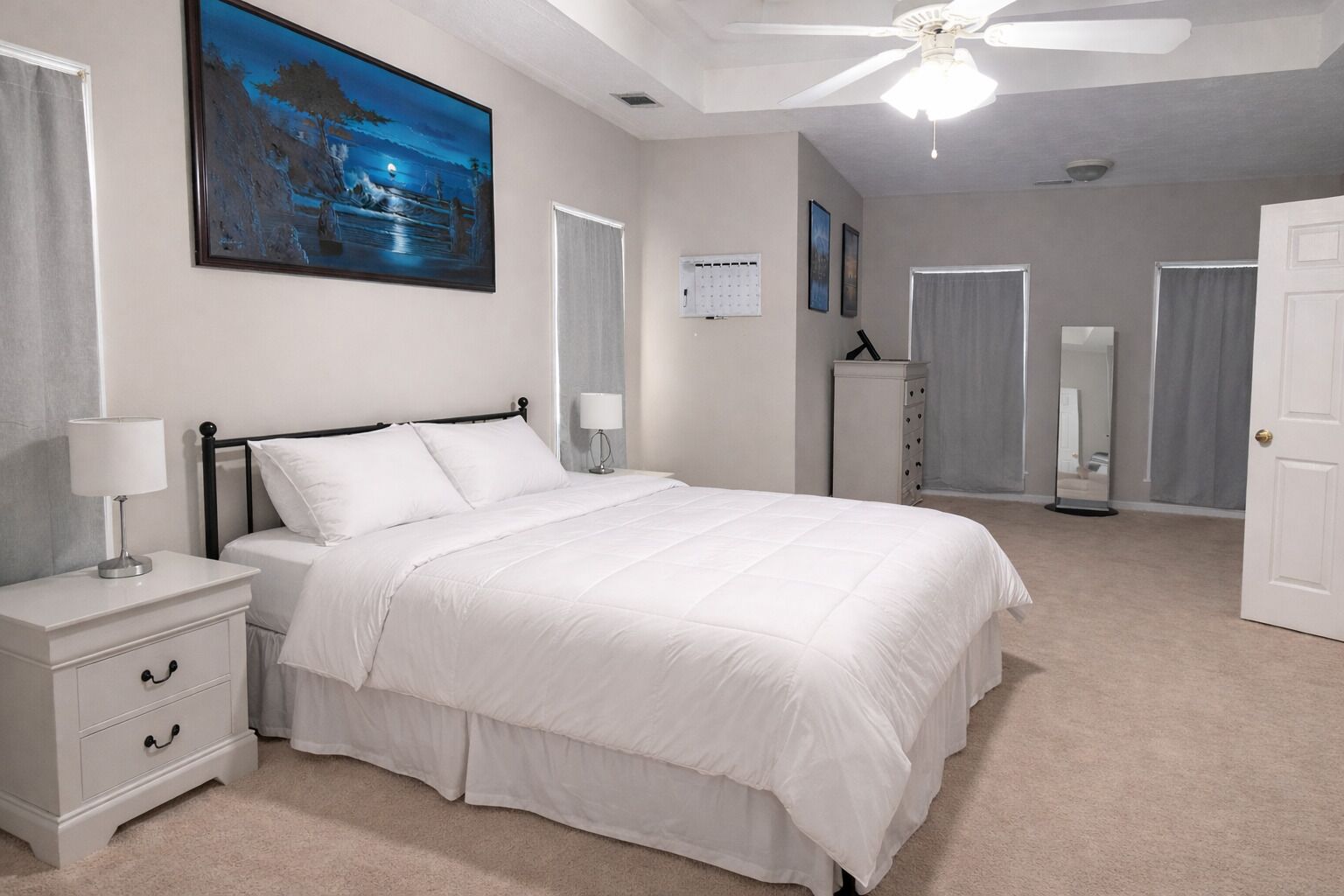 A bedroom with a bed, nightstands, a ceiling fan, a painting, a mirror, and a door.