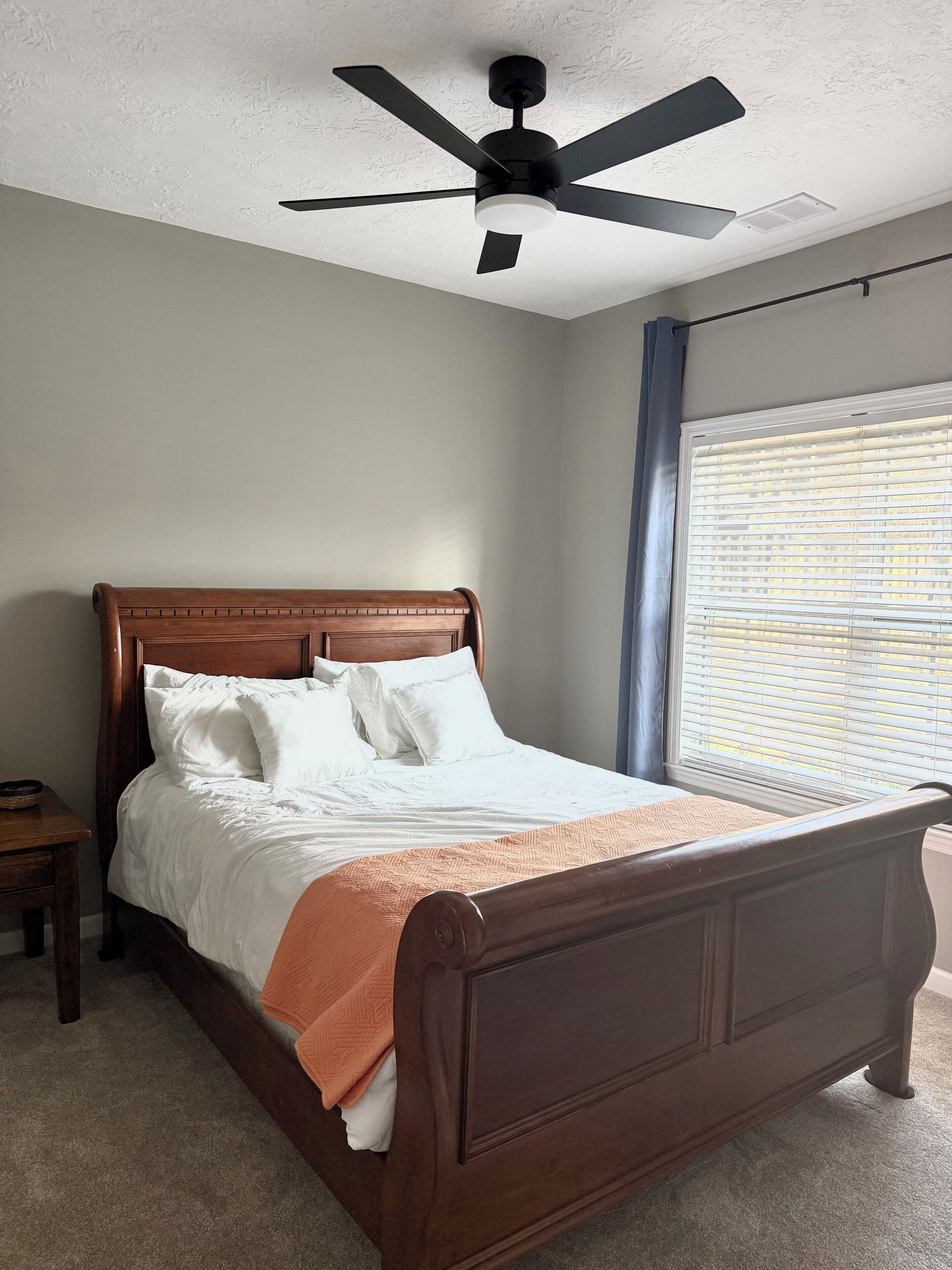 A bedroom with a wooden bed, a ceiling fan, a nightstand, and a window with blinds.