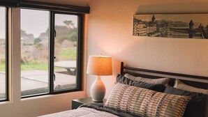 A bedroom with a bed, a window with a view, a lamp, and a painting on the wall. - Charming 2-bedroom house in Sea Ranch perfect for relaxing getaway (Sea Ranch)