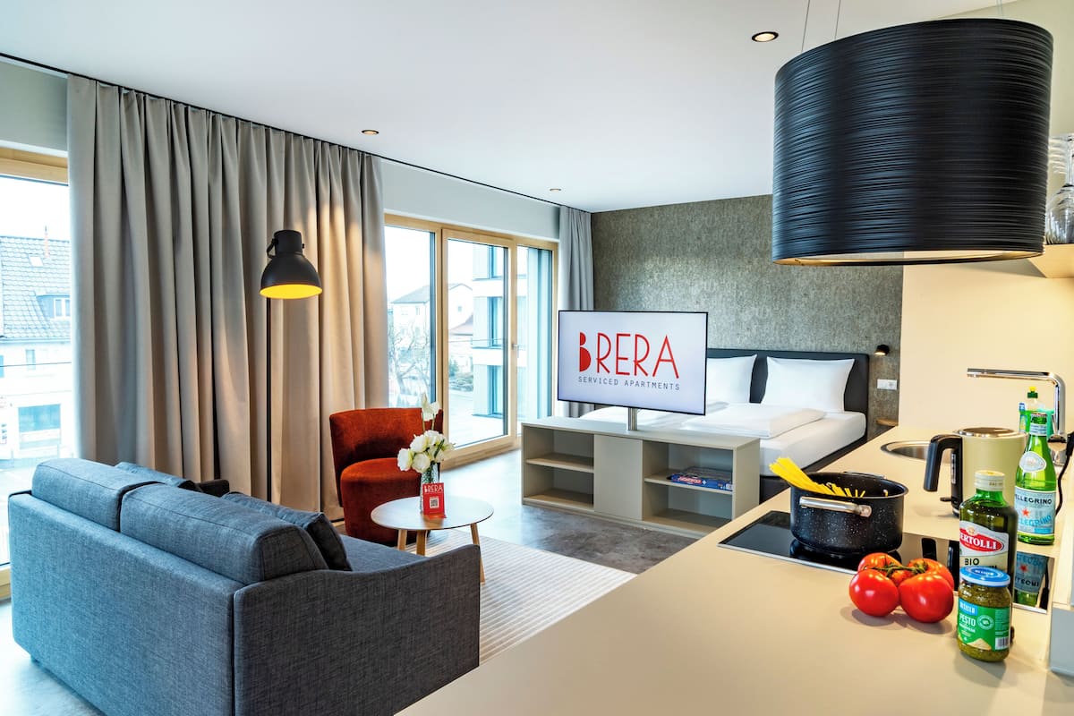 Ein modernes Wohnzimmer mit einer Couch, einem roten Sessel, einem kleinen Tisch mit einer Vase und einer Küchenzeile mit einem Topf, Tomaten und Flaschen.