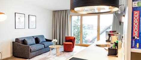 Een moderne woonkamer met een bank, een rode stoel, een kleine tafel met een vaas en een keukenhoek met een spoelbak en een Monopoly spel.