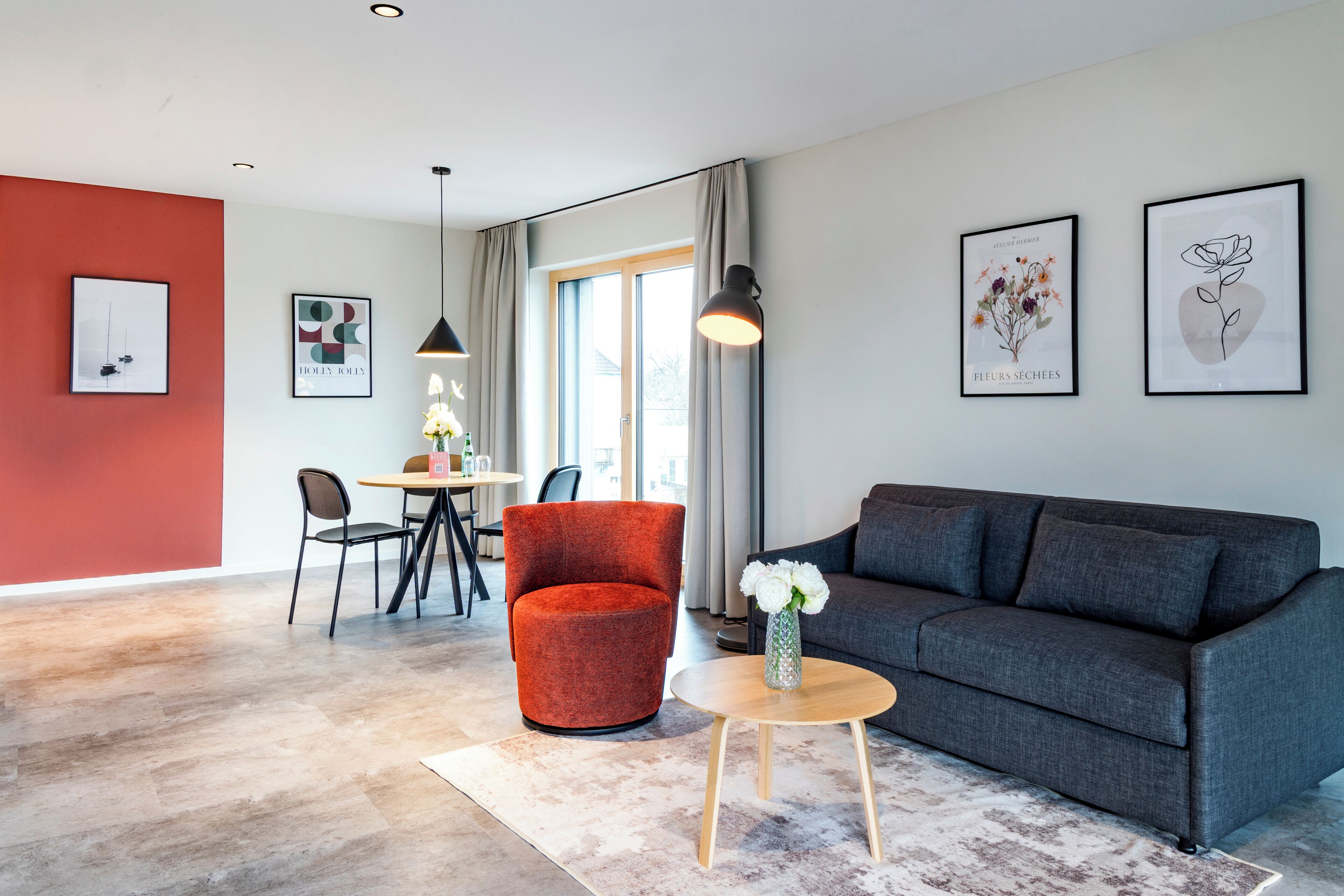 Una sala moderna con un sofá gris, un sillón rojo y una mesa redonda de madera con un jarrón de flores. Hay dos cuadros enmarcados en la pared, una zona de comedor con mesa y sillas, y un ventanal con cortinas.