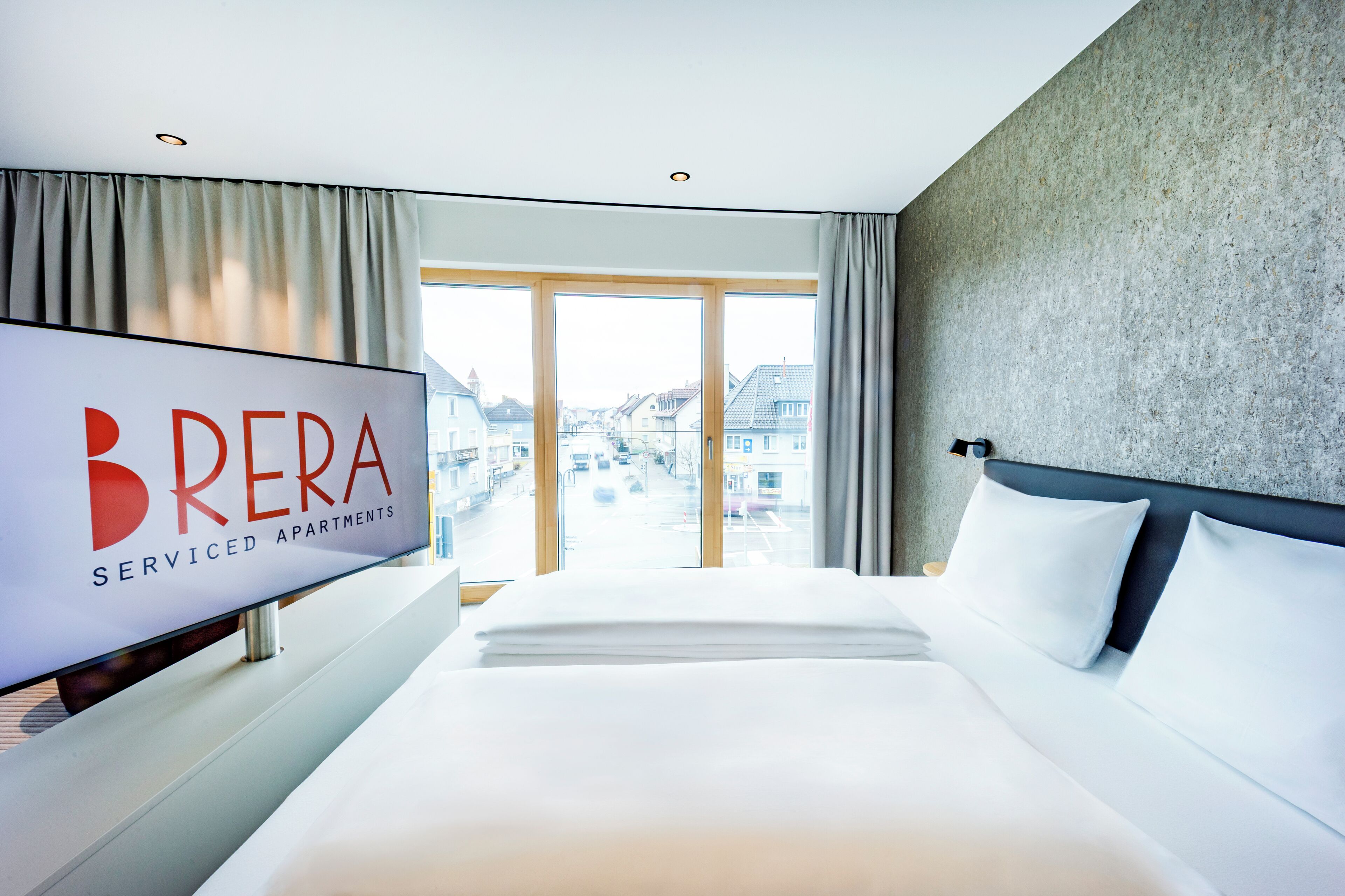 A hotel room with a large bed, a flat-screen TV, and a view of buildings through large windows.
